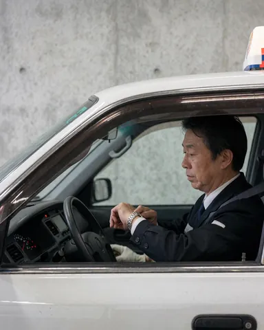 Un chauffeur de taxi assis dans son véhicule stationné consulte sa montre. L'éclairage intérieur et un mur en béton sont visibles en arrière-plan.