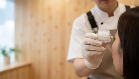 Professionnelle en gants blancs tenant un récipient de gel devant l'œil d'un client dans un salon de beauté.