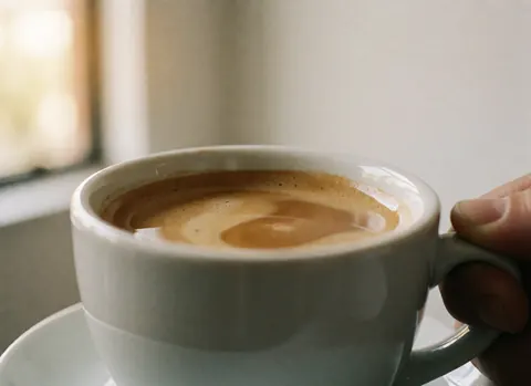Tasse blanche de café avec micro-mousse tenue par une main, fond blanc et lumière naturelle, idéal pour une cafétéria.