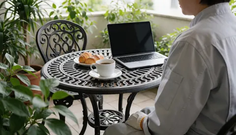 Une personne travaille sur une terrasse avec une table en fer forgé, un ordinateur portable, une tasse de café et des pâtisseries, idéal pour une maison d'hôtes.