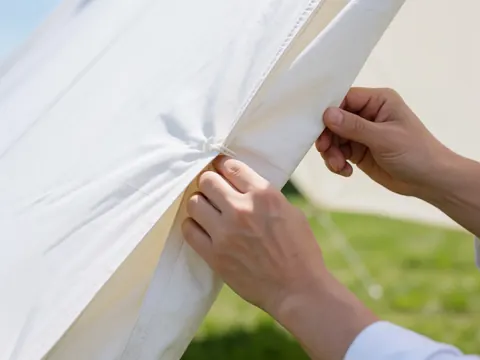 Des mains ajustent le tissu blanc d'une tente installée sur une pelouse verte en extérieur sous lumière naturelle.