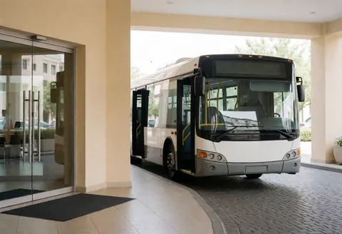 Autobus touristique blanc aux portes ouvertes, prêt à partir devant l'entrée d'un hotel moderne aux murs beiges et au sol en pierre.