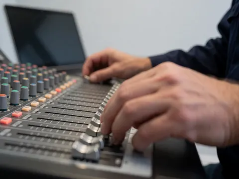 Gros plan sur des mains réglant les curseurs d'une table de mixage sonore, avec un ordinateur portable et un mur gris en arrière-plan, évoquant la production audiovisuelle.