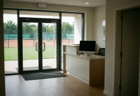 Entrée moderne d'un club de tennis avec réception, offrant une vue sur des courts en terre battue et en gazon à travers des portes vitrées.