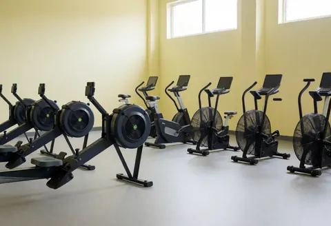 Espace d'entraînement avec bancs de rame et vélos stationnaires, murs jaunes clairs, lumière naturelle par fenêtres hautes, idéal pour un gymnase de CrossFit.