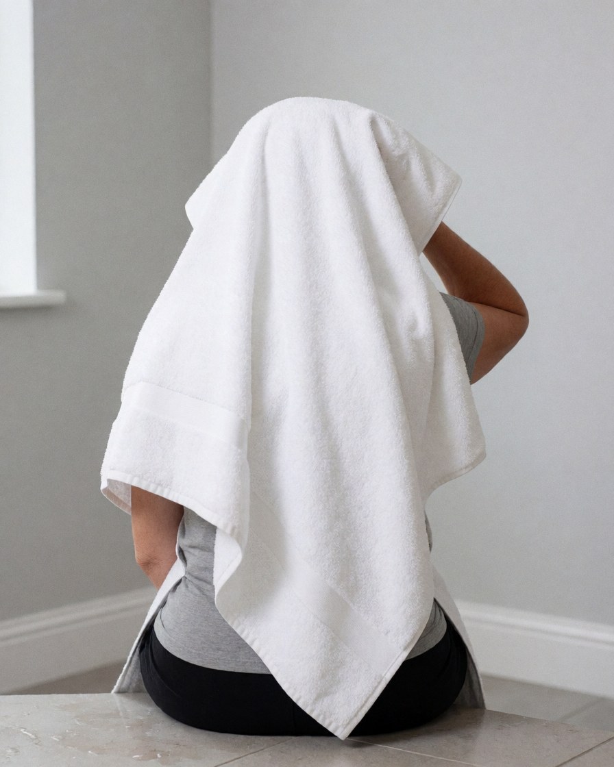 Une personne assise sur un sol carrelé avec des murs gris clair, séchant ses cheveux avec une serviette blanche, idéale pour une maison de vacances.