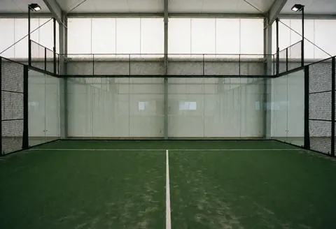 Court de tennis intérieur doté de murs vitrés et d'un sol vert, idéal pour les installateurs de terrains de tennis et de padel.