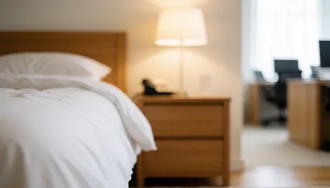 Lit avec draps blancs et table de chevet en bois dans une chambre d'aparthotel à la lumière chaude et aux murs blancs, idéale pour du texte publicitaire