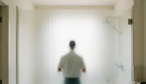 Un installateur pose un panneau de verre dépoli dans une salle de bain moderne aux murs blancs chauds, avec un design élégant et une lumière diffuse.
