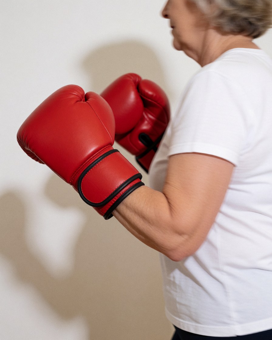 Une femme âgée en mouvement avec des gants de boxe rouges devant un mur blanc, idéale pour illustrer le fitness ou la défense personnelle féminine.