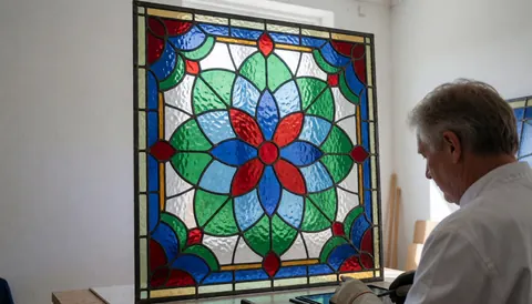 Un artisan réalise un panneau de vitrail à motif floral dans un atelier lumineux, avec du verre coloré et des cames de plomb visibles.