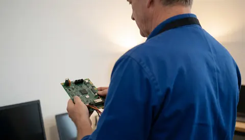 Un technicien en train de vérifier une carte de circuit dans un atelier avec un fond blanc et une lumière chaude pour la réparation de téléviseurs.