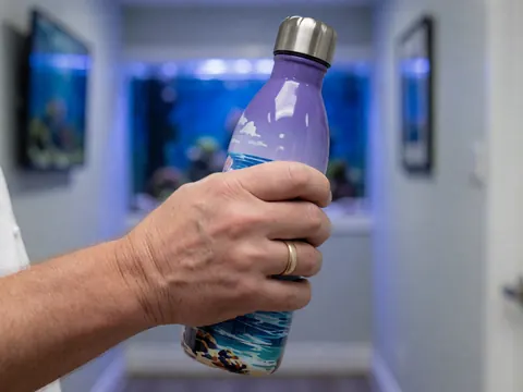 Une main tenant une bouteille décorée de motifs marins dans un couloir d'aquarium éclairé en bleu avec des murs gris.