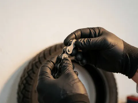Mains portant des gants noirs ajustant une chaîne de moto avec un pneu en arrière-plan dans un atelier à mur blanc et lumière chaude.