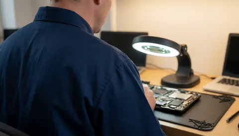 Un technicien en réparation de ordinateurs travaille sur une carte mère à l'aide d'une loupe de travail posée sur une table de travail anti-statique.