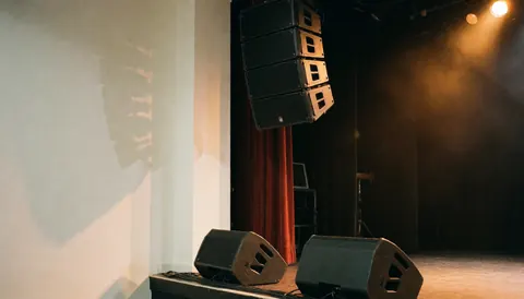 Scène de concert équipée d'une ligne de haut-parleurs suspendus et d'enceintes au sol, éclairage chaud et rideaux rouges, idéale pour des événements musicaux professionnels.