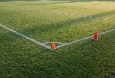 Terrain de football naturel avec lignes blanches, deux cônes orange près du coin et longues ombres de la lumière de l'aube, idéal pour entraîneur de football.