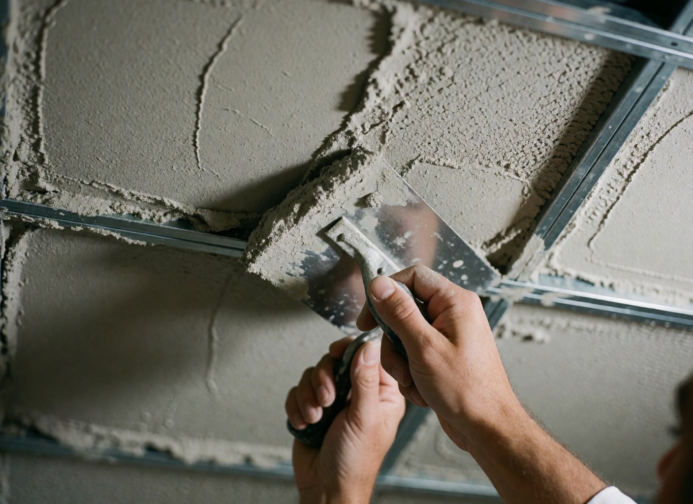 Gros plan d'une main utilisant une truelle pour appliquer du matériau gris sur une structure métallique, révélant une texture rugueuse sous un éclairage directionnel pour un professionnel du bâtiment.
