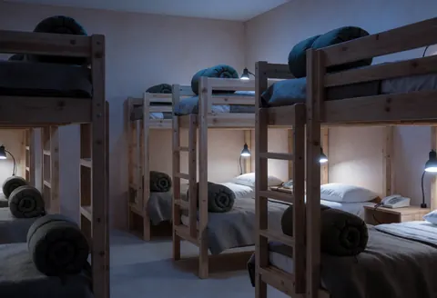 Chambre d'hébergement avec lits en bois, couvertures roulées, lumières de lecture et murs terracotta éclairés par une lumière bleue froide.