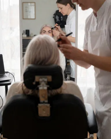 Une cliente aux cheveux clairs regarde son nouveau maquillage des sourcils dans un miroir pendant qu'une technicienne applique un produit dans un studio bien éclairé.
