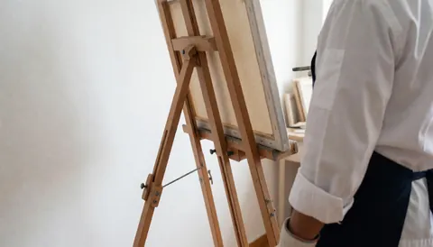 Chevalet de peinture avec une toile blanche vierge posée dessus, dans un studio aux murs blancs, idéal pour illustrer un cours de peinture.