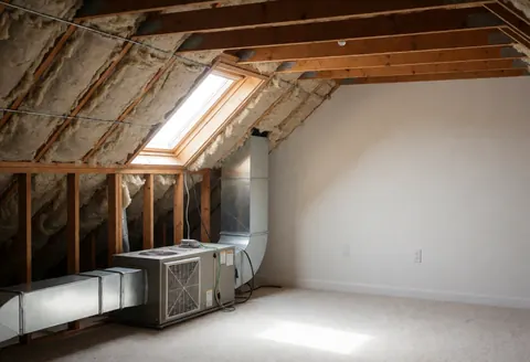 Vue intérieure d'un espace avec des conduits métalliques de chauffage et d'aération, des poutres en bois apparentes et une fenêtre de toit laissant entrer la lumière naturelle.