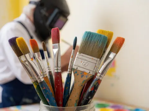 Des pinceaux colorés dans un pot avec un artiste travaillant en arrière-plan dans un espace aux murs chauds et à la lumière vive.