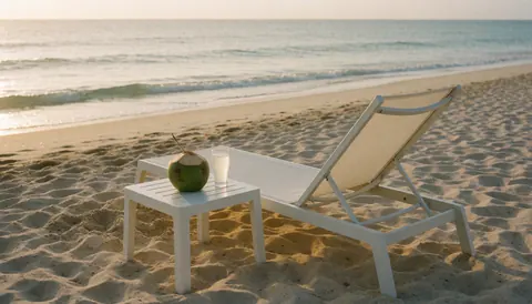 Chaise de plage blanche posée sur le sable avec un coco et un verre d'eau de coco, des vagues de mer en arrière-plan, ambiance idéale pour un club de plage.
