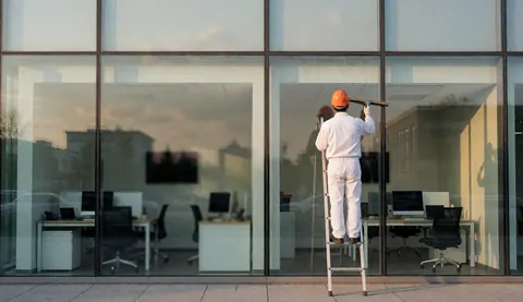 Un ouvrier en uniforme blanc et casque orange utilise une raclette sur une fenêtre de bureau, avec un reflet de coucher de soleil sur la vitre.