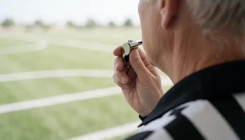 Vue arrière d'un arbitre de football portant un uniforme rayé noir et blanc, soufflant dans son sifflet sur un terrain de sport en plein jour.