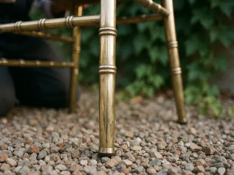 Chaise en bronze aux pieds fins posée sur du gravier, entourée de feuilles vertes et de reflets lumineux dorés, idéale pour la location de mobilier d'événement.