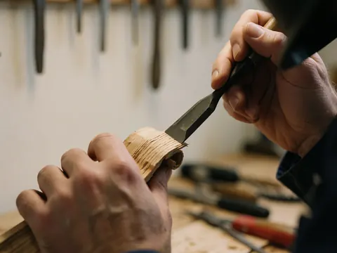 Gros plan d'une main utilisant un ciseau sur du bois de chêne, avec des outils suspendus en arrière-plan flou dans un atelier de menuiserie.