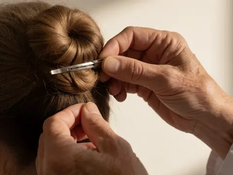 Gros plan de mains ajustant une épingle à cheveux métallique dans un chignon brun sur fond blanc, pour un instructeur de danse.