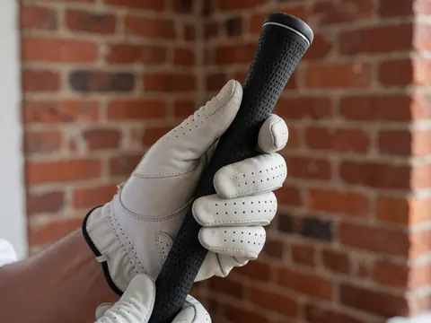 Gros plan d'une main portant un gant de golf en cuir perforé tenant fermement une poignée noire, avec un mur de briques rouges en arrière-plan.