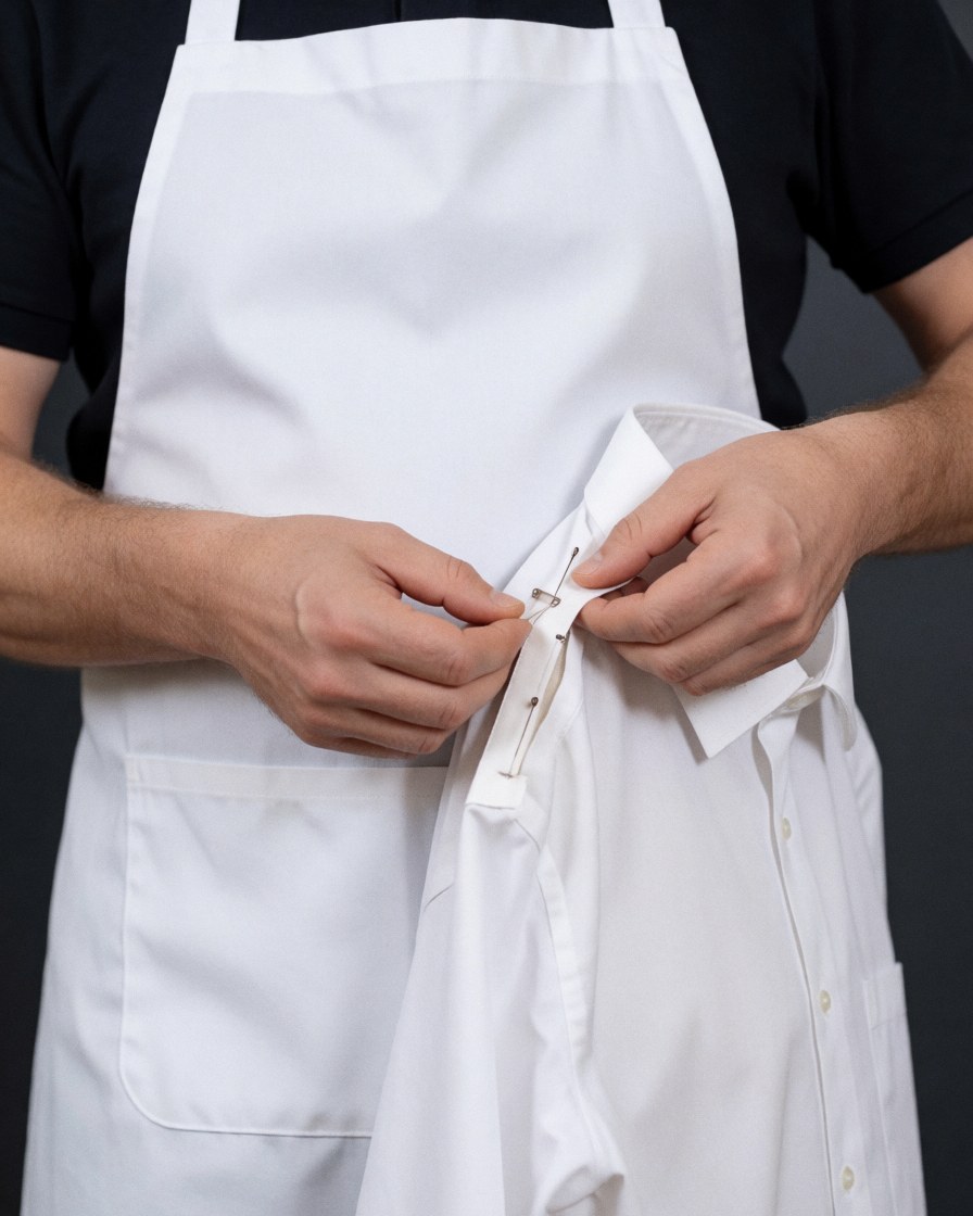 Mains d'un couturier épinglant le col d'une chemise blanche avec un tablier blanc sur fond gris foncé pour la réparation de vêtements.