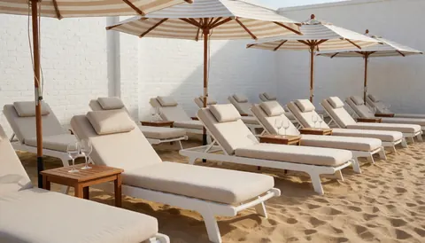 Transats blancs avec coussins beiges sous des parasols rayés sur une plage avec un mur de briques blanches et du sable doré, idéal pour un club de plage.