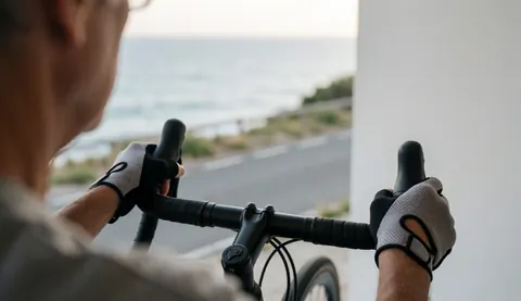 Gros plan sur des mains portant des gants de cyclisme tenant le guidon d'un vélo sur une route côtière au coucher du soleil avec vue sur la mer.