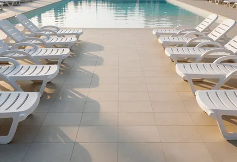Des chaises de plage blanches sont disposées sur un sol carrelé beige, face à une piscine aux eaux claires, baignée par une douce lumière matinale.