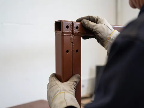 Une main portant un gant de travail ajuste une structure métallique peinte en rouge avec des trous de vis dans un atelier à mur blanc.
