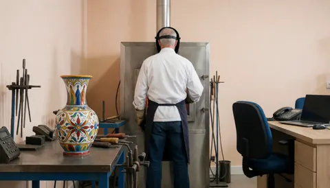Un artisan travaille au four à verre, un vase décoratif repose sur une table, des outils et des murs beiges sont visibles dans l'atelier.