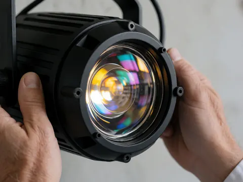 Objectif de projecteur de scène montrant des reflets de gels chromatiques, mains ajustant l'équipement sur fond de mur en béton lisse dans un centre de spectacles.
