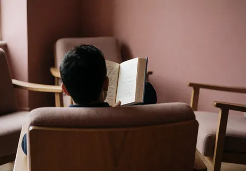 Une personne assise lisant un livre ouvert dans un intérieur calme avec des murs roses et des chaises en bois, évoquant une ambiance studieuse.