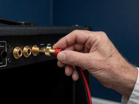 Une main adulte branche un câble Speakon doré à isolation rouge et noire sur un amplificateur noir, contre un mur bleu marine, pour des événements sonores.