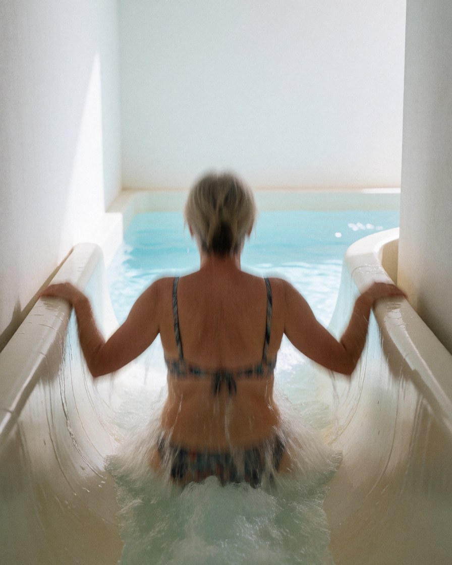 Un adulte descend un toboggan dans une piscine extérieure avec de l'eau qui éclabousse et des murs blancs en arrière-plan.