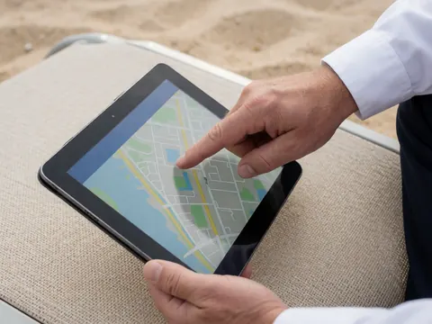 Une main effleure une carte numérique affichée sur une tablette noire, posée sur une transat beige en bord de plage, évoquant la réservation d'un club de plage sous un éclairage naturel.