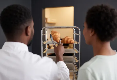 Une cliente prend un croissant sur une étagère métallique dans la kitchenette d'un apart-hotel aux murs gris et à l'éclairage chaleureux.