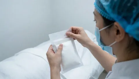 Un thérapeute en tenue de protection ouvre un paquet de compresses blanches sur une table de soin dans un cabinet de soins de la peau.