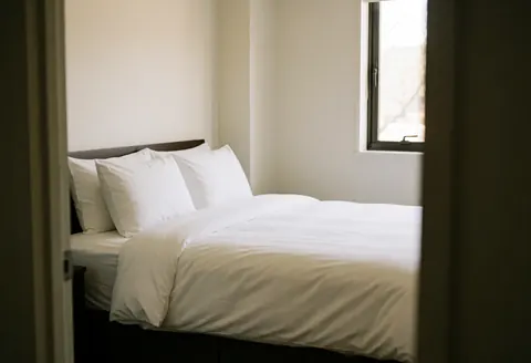 Lit avec draps blancs et oreillers dans une chambre propre aux murs blancs chaudres et à la lumière naturelle par la fenêtre, idéal pour une maison de vacances.
