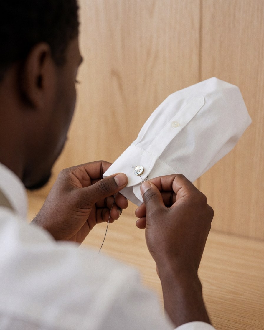 Gros plan d'une main experte en train de recoudre un bouton en nacre sur une chemise blanche, sur un fond en bois clair, illustrant le service de réparation de vêtements.