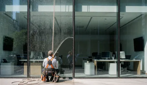 Un ouvrier en harnais et corde nettoie une grande vitre d'un immeuble de bureaux moderne, révélant l'intérieur avec des bureaux et des ordinateurs.
